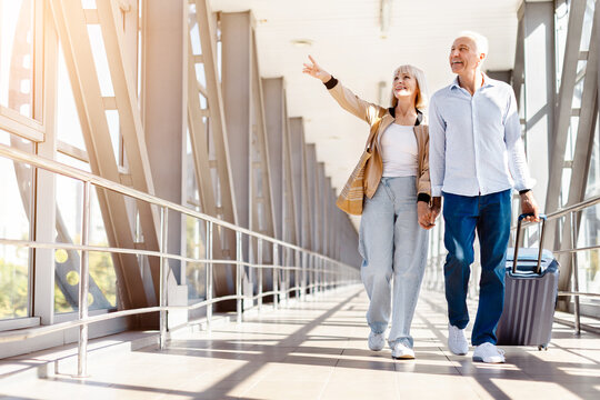 ature couple walking in airport terminal with luggage, woman pointing ahead while smiling, bright corridor scene representing shared travel experience and future journey direction
