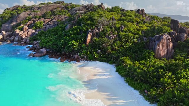 Turquoise ocean waves wash the white sand beach in the Seychelles. Island aerial captures jungle trees and granite boulders bordering the coast of the Seychelles