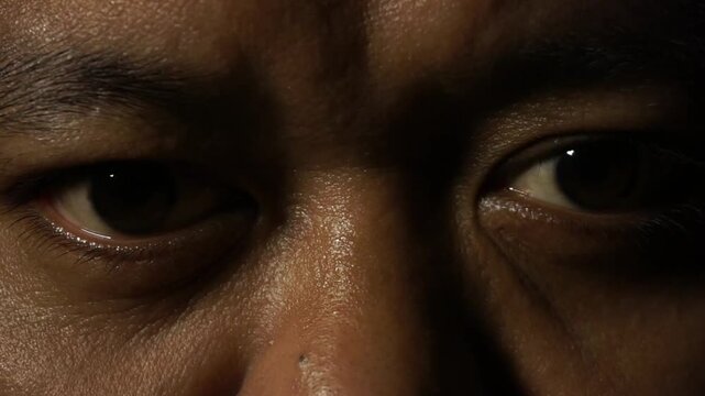 Warm flashing light reveals a close up of a sweating male face, his dark eyes staring intensely forward out of the deep shadows.