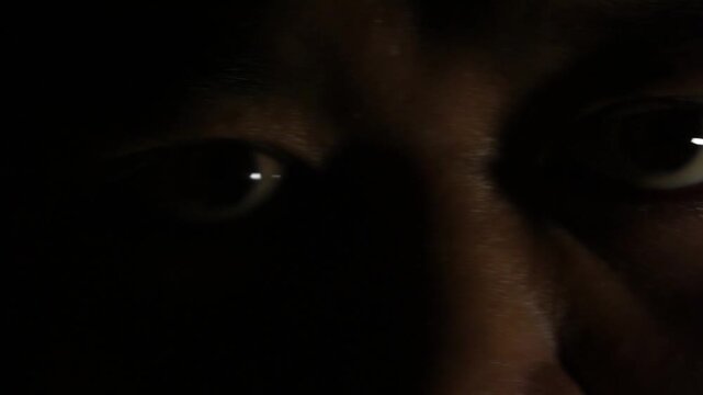 Flashing warm light reveals the intense stare of a male subject in deep darkness, his eyes shifting focus and blinking rapidly.