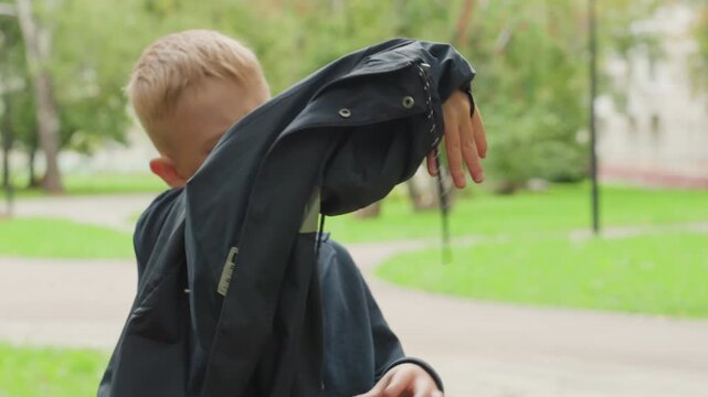 White boy struggling with jacket near park path, fumbling with sleeve and zipper, annoyed expression and determined effort, candid closeups capture frustration and small victory while preparing to go