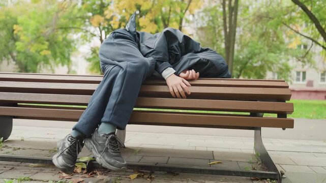 Tired student resting on bench under trees, denim jeans and dark jacket, hooded slouch, autumn leaves on ground, quiet park scene, reflective mood as student prepares for return to school