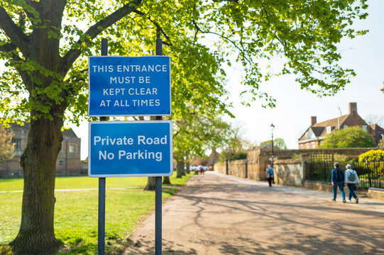 Large blue No Parking and Private Road sign seen in the historic cathedral city of Ely, UK. A public park is on the left, with passers by seen walking on the right.
