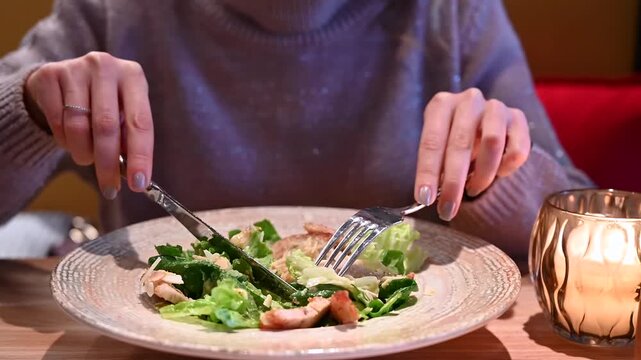 Woman uses knife and fork to eat caesar salad with chicken at restaurant table.