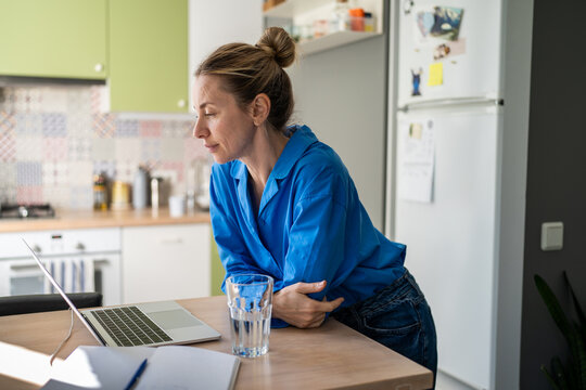 Interested calm mature woman watching media online on laptop, enjoying spare time at home during work break. Positive female browsing online on computer in kitchen, looking through viral content.