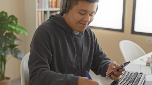 Man latin hispanic with over ear headphones holds smartphone and writes notes with pen at a desk in an office building while seated and checking the screen; concentration productivity.