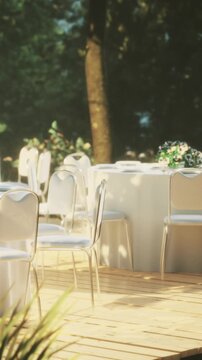 A round table is set up outdoors under trees. It has a floral centerpiece and several empty chairs around it. The setting is for a gathering or celebration in the daytime.