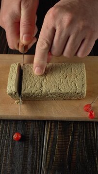 Hands carefully cut a block of halva on a wooden cutting board, with red berries scattered around, emphasizing the process of making the dessert