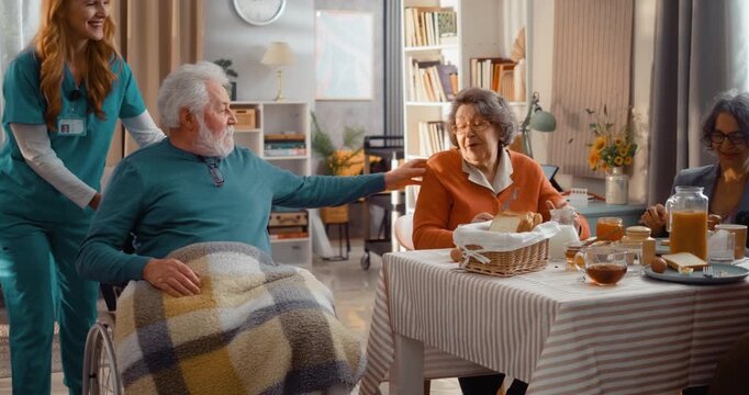 Kind female caregiver helping elderly man in wheelchair join group breakfast. Warm moment of care, accessibility, dignity and social interaction in assisted living setting.