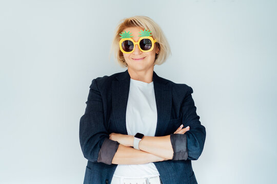 Middle age woman with crossed arms in blazer and white outfit posing in pineapple sunglasses over isolated white background, smiling to the camera. Relaxed business woman.