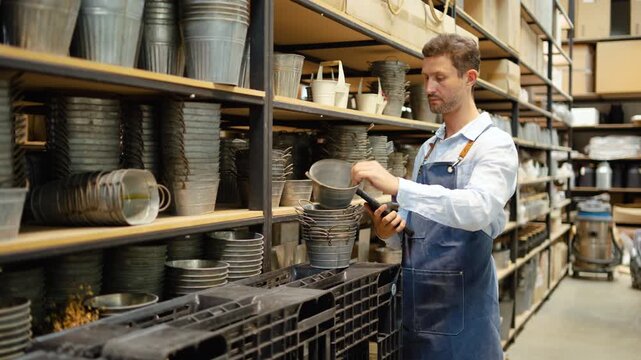 Male entrepreneur in apron using digital tablet to manage inventory of galvanized metal pots in warehouse. Professional small business owner conducting stock check and retail store management.