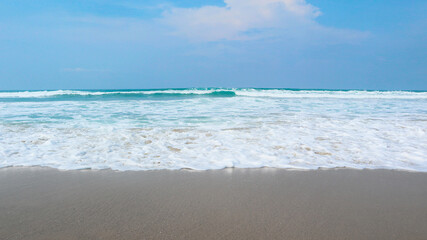 Pristine beach where vibrant turquoise ocean waves break into a thick layer of white sea foam. The frothy water recedes over smooth, wet, grey sand under a clear blue sky with soft, scattered clouds. © gusense