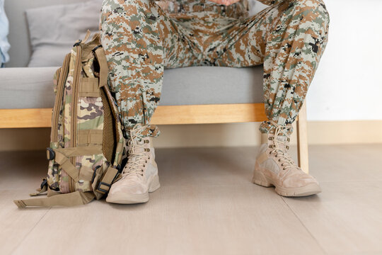Military soldier legs in camouflage uniform with tactical boots and backpack sitting on sofa at home, army service concept, returning from duty, rest, transition to civilian life.