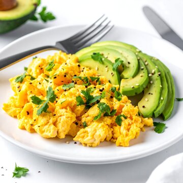 Breakfast dish on a white plate. Scrambled eggs are piled near sliced avocado, sprinkled with herbs and seeds. Silverware is beside