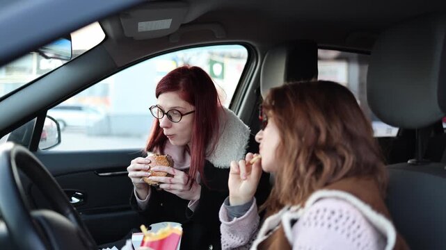 Two women eat burgers and fries inside parked car enjoying casual break together relaxed mood friendship moment everyday lifestyle fast food snack in vehicle interior
