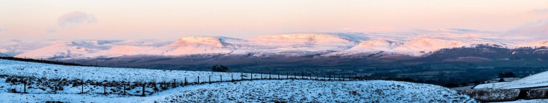 A panoramic view of snow-covered hills and valleys bathed in the soft, pinkish light of sunrise or sunset, creating a serene winter landscape.