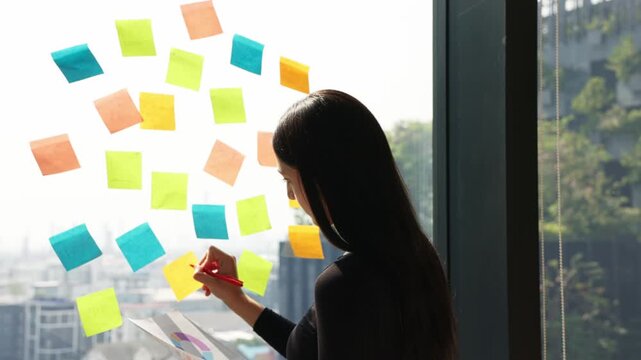 Young professional organizing ideas on colorful sticky notes while reviewing business chart near office window during planning session.