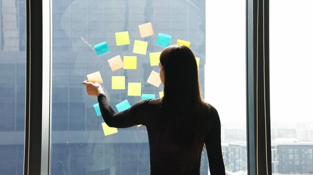 Young professional organizing ideas on colorful sticky notes while reviewing business chart near office window during planning session.