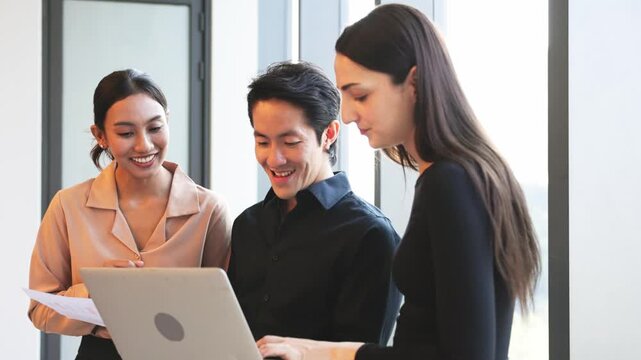 Three business professionals standing together in modern office reviewing charts and information on laptop during collaborative work discussion.