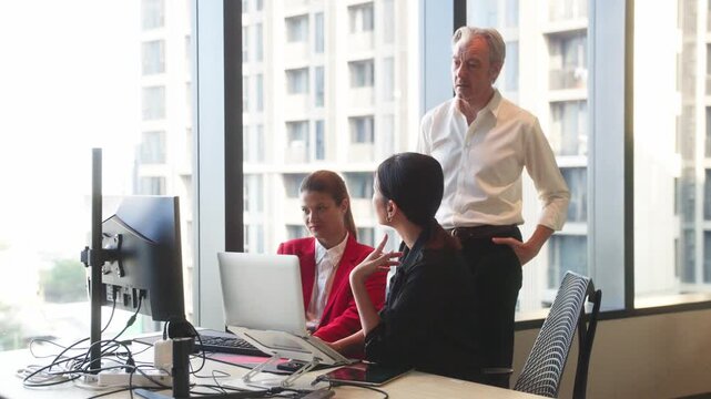 Three business professionals discussing project strategy around laptop in modern office meeting space with plants and collaborative atmosphere.