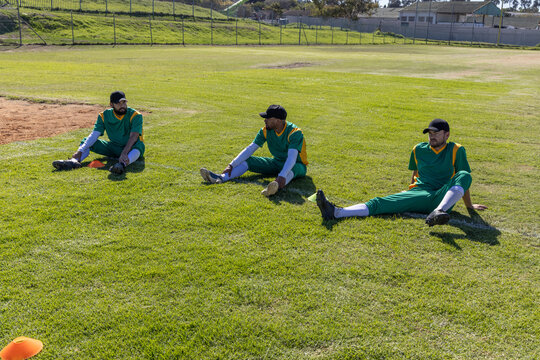 Male players sitting on grass by infield with orange cones wearing green-yellow jerseys black caps