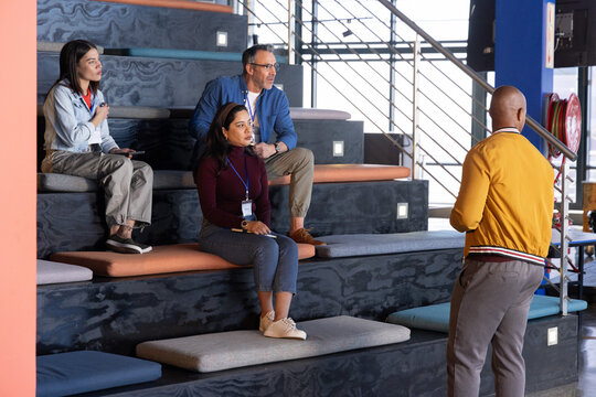 Diverse coworkers sitting on tiered benches in coworking space, wearing lanyards, facing presenter