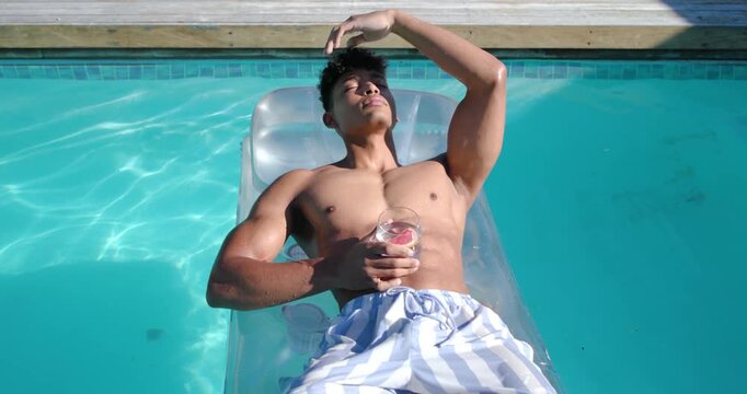 African American man mid-20s lounging on pool float in striped trunks with pink drink shading eyes
