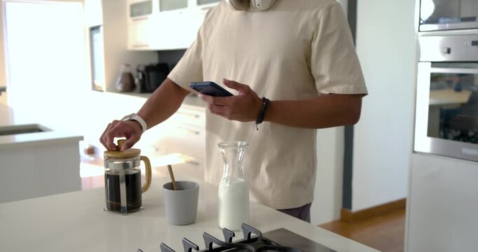 African American man checking phone, pressing plunger on French press, pouring coffee in kitchen