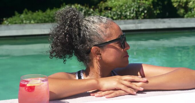 Senior African woman noticing camera turning to face smiling in swimsuit lifting pink drink at pool