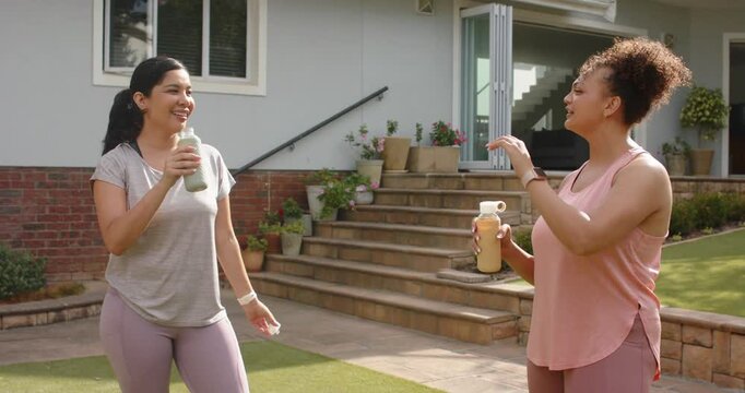 Diverse female friends chatting on patio, checking smartwatch, clinking reusable bottles for toast