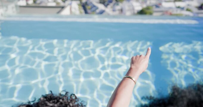 Diverse female friends pointing at horizon, relaxing in infinity pool, clinking drinks, in swimwear