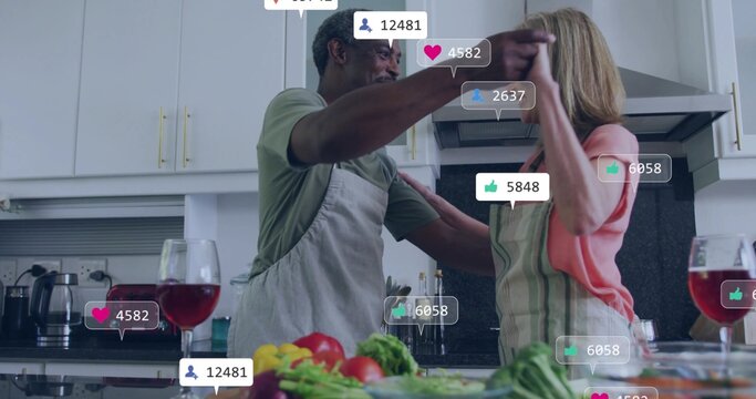 Dancing mature pair preparing meal at kitchen island, wearing aprons with produce, wine, reactions
