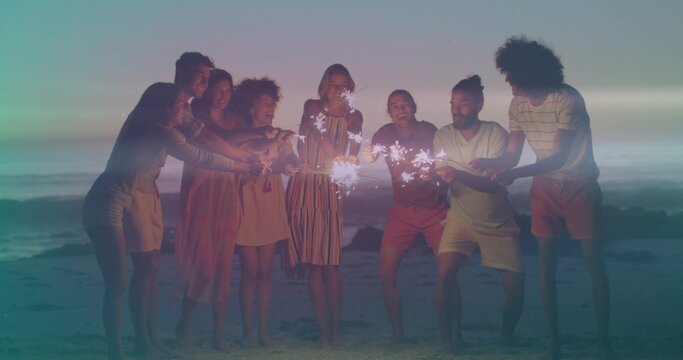 Gathering 8 adults in summer wear laughing, holding sparklers barefoot on sand at dusk, gentle surf