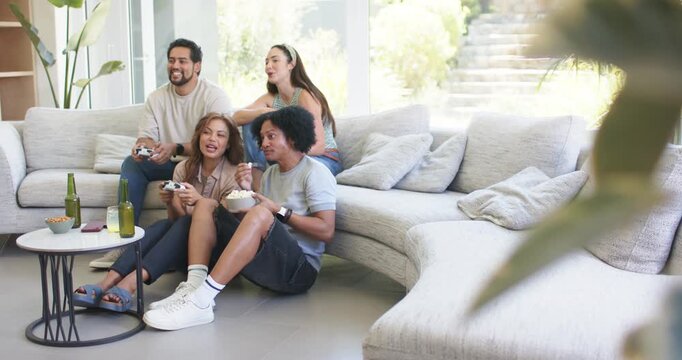Diverse friends grabbing controllers and snack bowls, playing game together and laughing at home