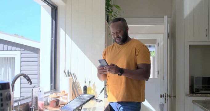 Mid-adult man in orange tee scrolling phone in kitchen charging on solar panel, reading texts