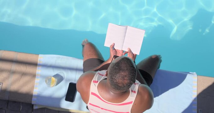 Mid-adult African man striped tank sitting poolside reading notebook, dipping feet to cool, phone