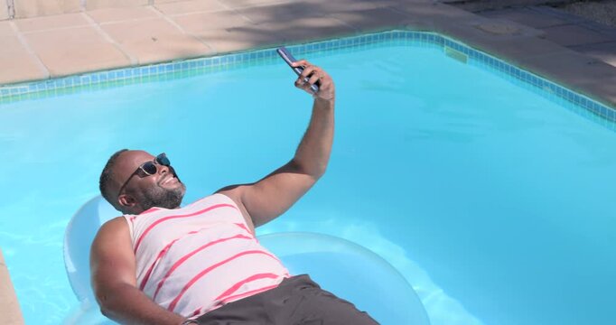 African American man in striped tank floating on float in home pool holding phone checking messages