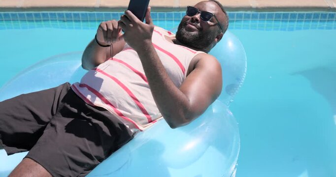African American man in striped tank reclining on pool float glancing at phone framing selfie