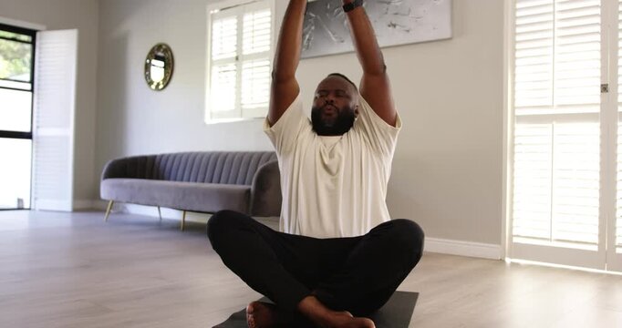 Breathing African man in white tee raising arms bringing palms to chest meditating on yoga mat
