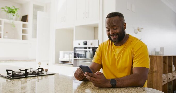 African man wearing yellow tee sitting at kitchen island reading texts on phone and smiling