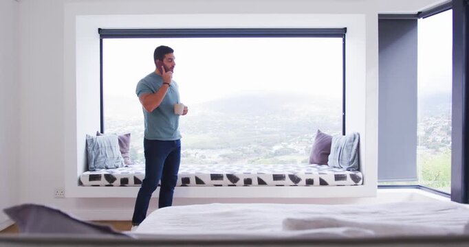 Adult male in 30s holding phone during call and mug, pacing past bed, pausing, stopping centered