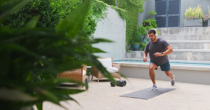 Man in his 30s stepping onto exercise mat, jogging, doing reverse lunges on patio for fitness