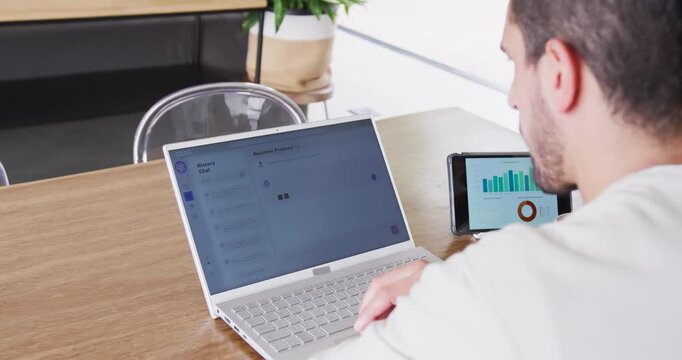 Adult man responding to smartphone chart, typing on laptop at wooden table, copy space