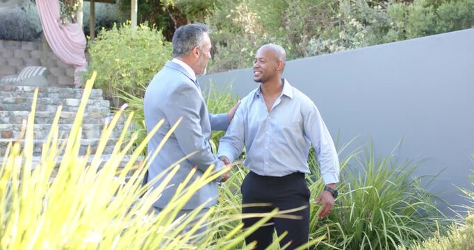 Diverse male coworkers coming, greeting, shaking hands, shifting to arm hold in garden with suit