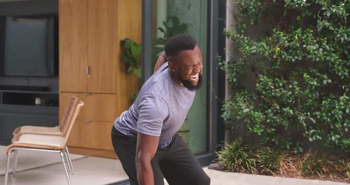 Man wearing gray tee, pants reaching for item at sliding door on patio, clutching lower back