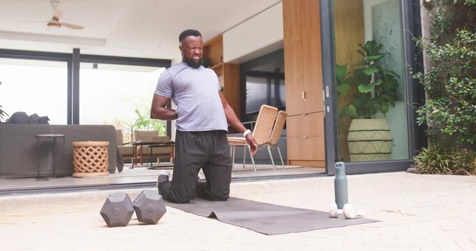 Man kneeling on yoga mat at patio, reaching back and bracing chair, seeking deeper chest stretch