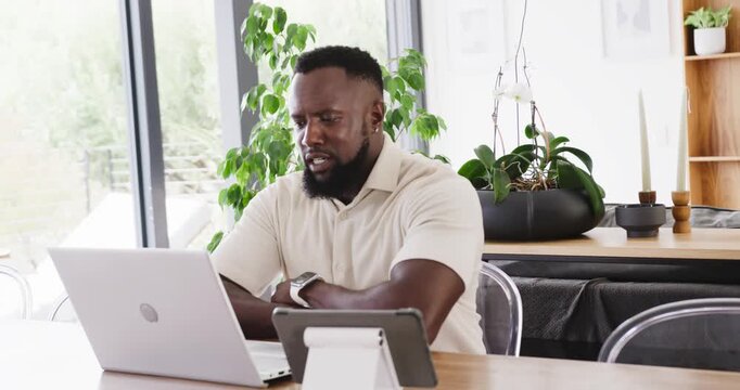 Man leaning forward typing on laptop in home with tablet, checking smartwatch, tech troubleshooting