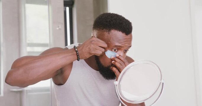 African American man taking blue under-eye patch leaning toward mirror smoothing patch for skincare