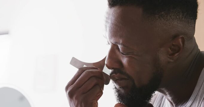 African American man in tank top lifting shaping tool and adjusting moustache line at vanity mirror