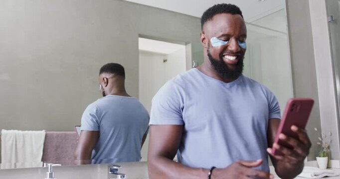 Man wearing under-eye gel patches at bathroom sink, tapping smartphone after text, laughing